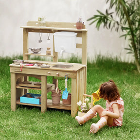 A young girl in pink boots enjoys hours of sensory play on green grass beside the Tiny Land® Mud Kitchen - DuoPlay Outdoor Play Kitchen for Kids, featuring utensils, a sink, and potted plants amidst lush garden greenery.