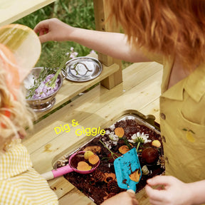 Two kids enjoy sensory play outdoors at the Tiny Land® Mud Kitchen - DuoPlay, using small shovels to dig in dirt, orange slices, and flowers. One wears yellow, matching the "Dig & Giggle" text. Outdoor fun with Tiny Land!.