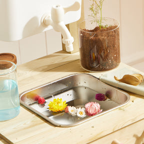 The Tiny Land® Mud Kitchen - DuoPlay Outdoor Play Kitchen for Kids features a metal sink in a light wooden counter, perfect for sensory play with water and flowers, plus nearby accessories like a glass, cloth, dried leaf, and potted plant.