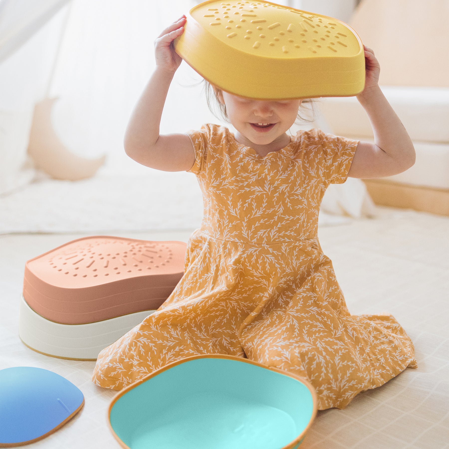 A young child in a yellow patterned dress sits on the floor with an upside-down yellow colander on their head. Tiny Land® Stepping Stones - Beach Vacation by Tiny Land are scattered around, perfect for obstacle course fun and motor skill development.|Beach Vacation