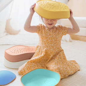 A young child in a yellow patterned dress sits on the floor with an upside-down yellow colander on their head. Tiny Land® Stepping Stones - Beach Vacation by Tiny Land are scattered around, perfect for obstacle course fun and motor skill development.|Beach Vacation