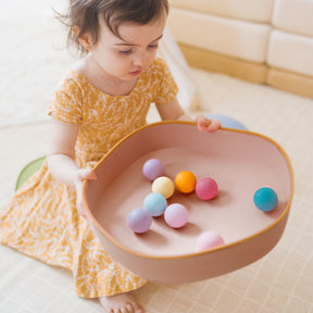 A young child in a yellow dress sits indoors on a light floor, holding a large pink bowl of colorful balls—perfect for developing motor skills after playing on the Tiny Land® Stepping Stones - Beach Vacation by Tiny Land.|Beach Vacation