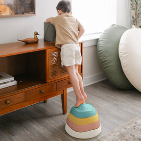 A young child stands on Tiny Land® Stepping Stones - Beach Vacation by Tiny Land, reaching for an object on a console table in a bright living room—ideal for building motor skills through this fun obstacle course.|Beach Vacation