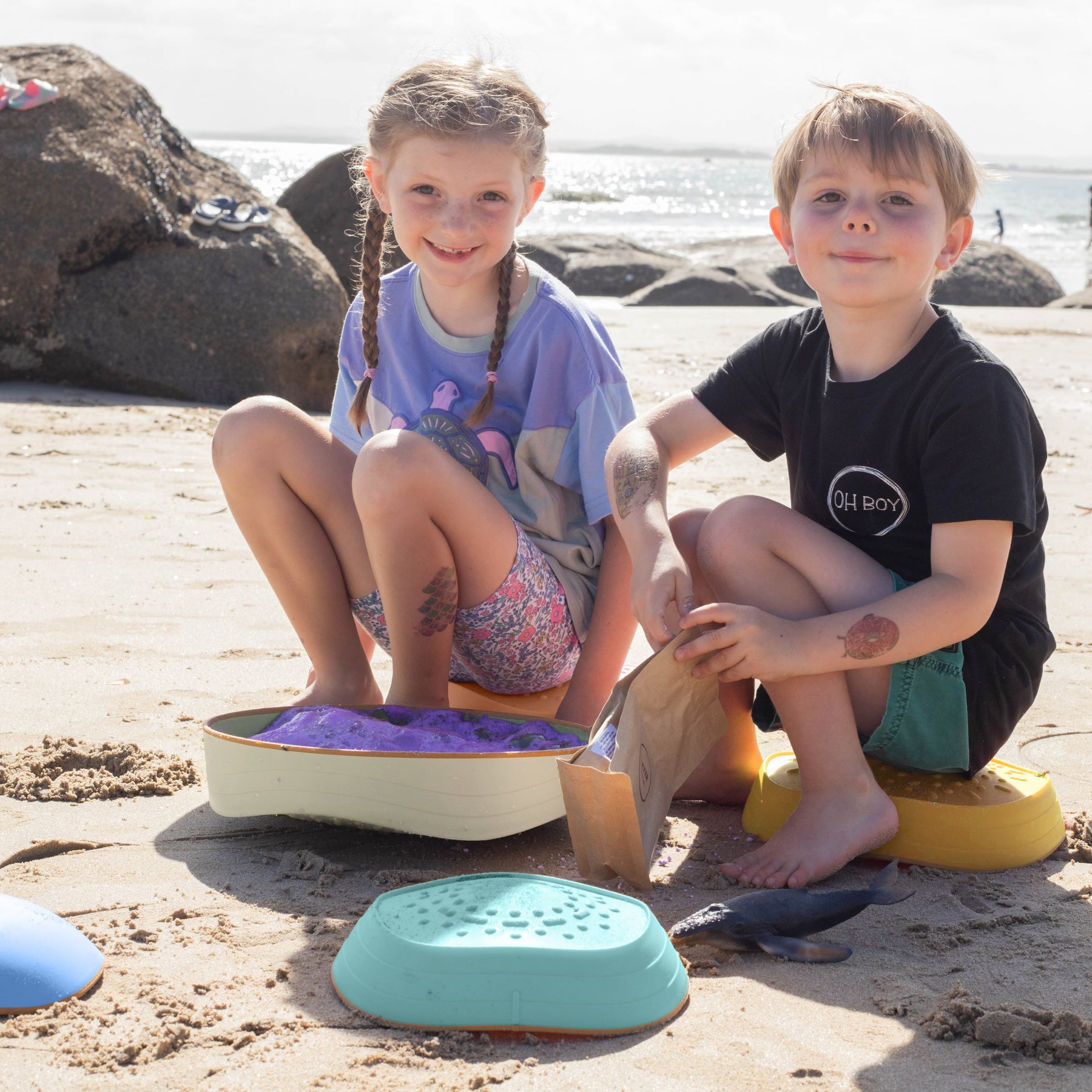 Two young children smile and play on the beach with sand toys and Tiny Land® Stepping Stones - Beach Vacation, building motor skills as waves sparkle behind them under a sunny sky.|Beach Vacation