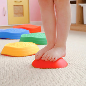 A child stands barefoot on a red Tiny Land® Stepping Stone - Rainbow Color by Tiny Land, with more rainbow stones lined up behind on a carpeted playroom floor, creating a fun obstacle course to enhance motor skills.|Rainbow Color