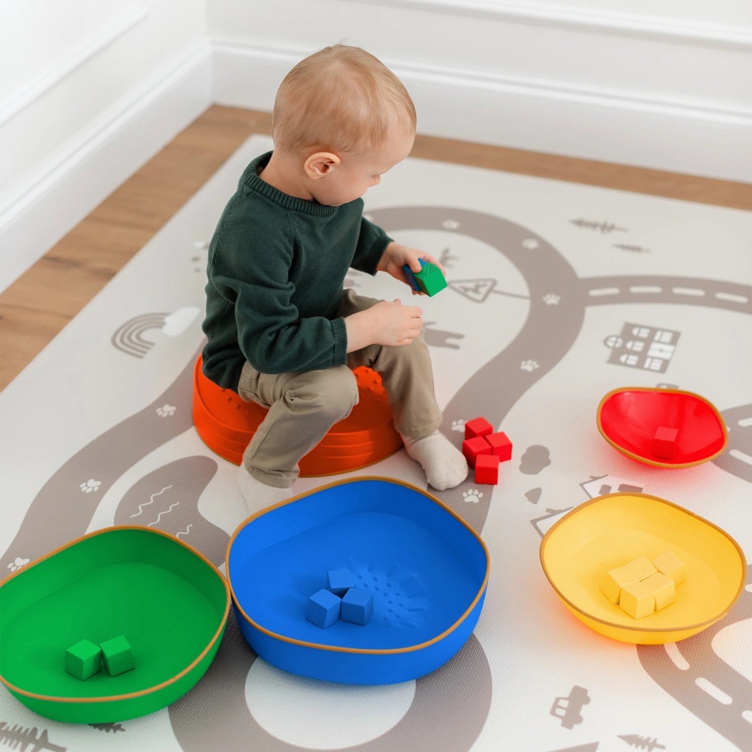 A toddler sits on an orange cushion, sorting colorful blocks into green, blue, red, and yellow bowls on a playmat with roads and buildings, building motor skills like with Tiny Land® Stepping Stones - Rainbow Color by Tiny Land.|Rainbow Color