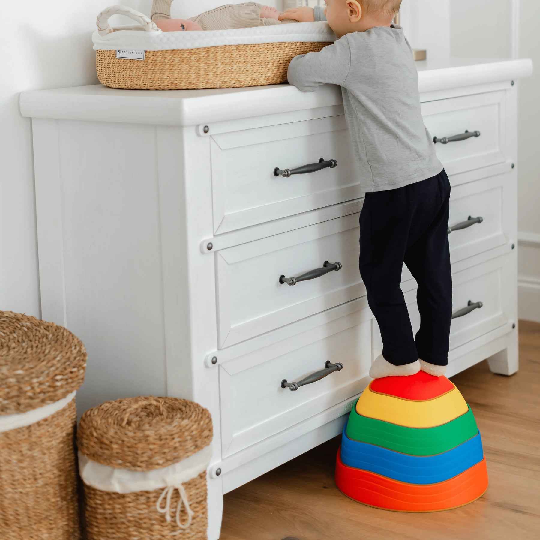 A toddler uses Tiny Land® Stepping Stones - Rainbow Color by Tiny Land to reach a changing table with a baby in a basket, while two woven baskets sit beside a white dresser, promoting balance and motor skill growth.|Rainbow Color