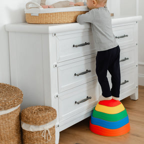 A toddler uses Tiny Land® Stepping Stones - Rainbow Color by Tiny Land to reach a changing table with a baby in a basket, while two woven baskets sit beside a white dresser, promoting balance and motor skill growth.|Rainbow Color