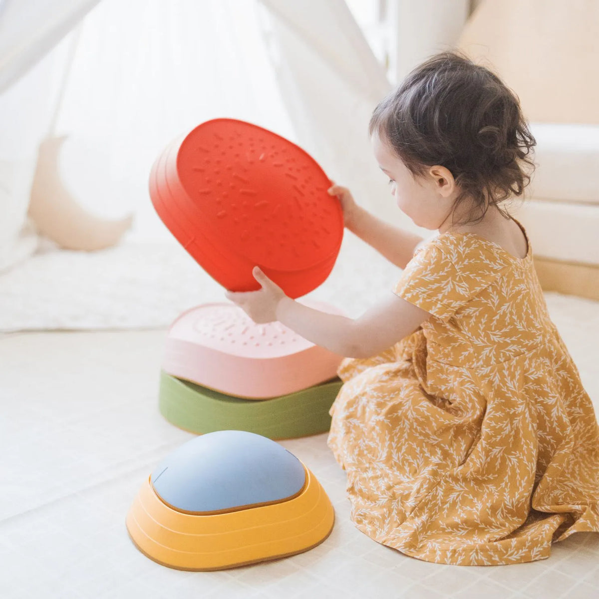 A young child in a yellow dress sits on the floor, stacking Tiny Land® Stepping Stones - Vintage Color by Tiny Land in a bright, cozy room—building motor skills for a playful obstacle course.|Vintage Color