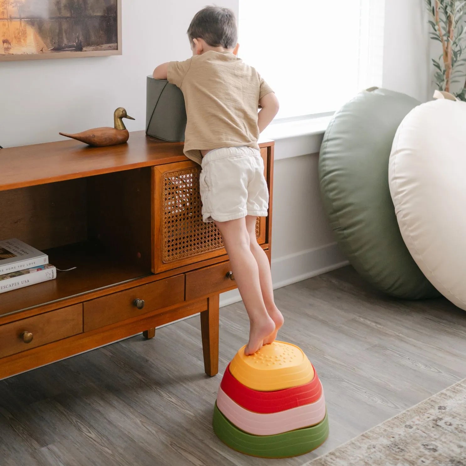 A young boy uses Tiny Land® Stepping Stones - Vintage Color from Tiny Land to reach a gray box atop a wooden cabinet, enhancing his motor skills in a bright, modern living room.|Vintage Color
