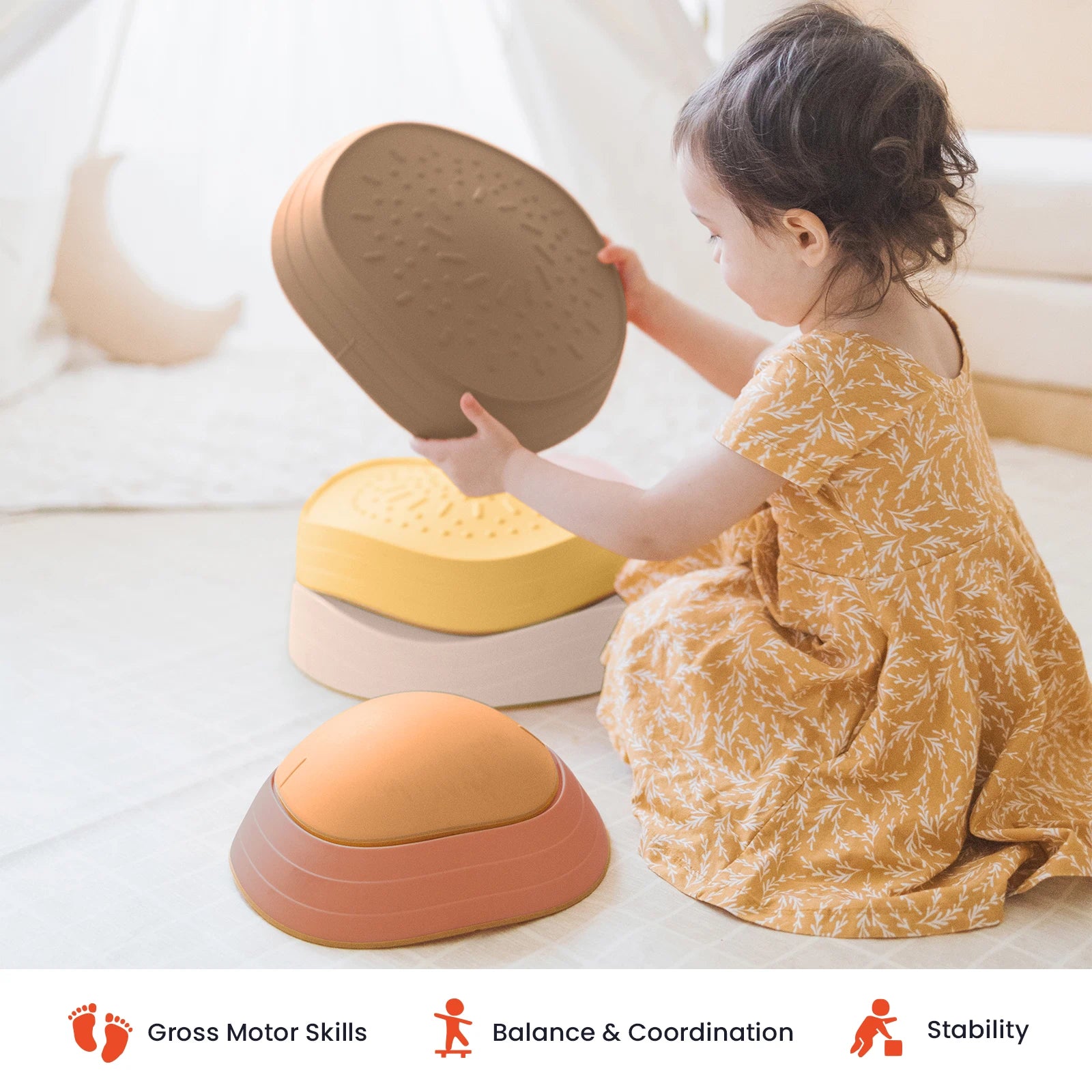 A young child in a yellow patterned dress stacks Tiny Land® Stepping Stones - Sunset Hues on a soft surface. Icons and text at the bottom highlight benefits: gross motor skills, balance & coordination, and stability—making these Tiny Land stepping stones perfect for creating an engaging obstacle course.