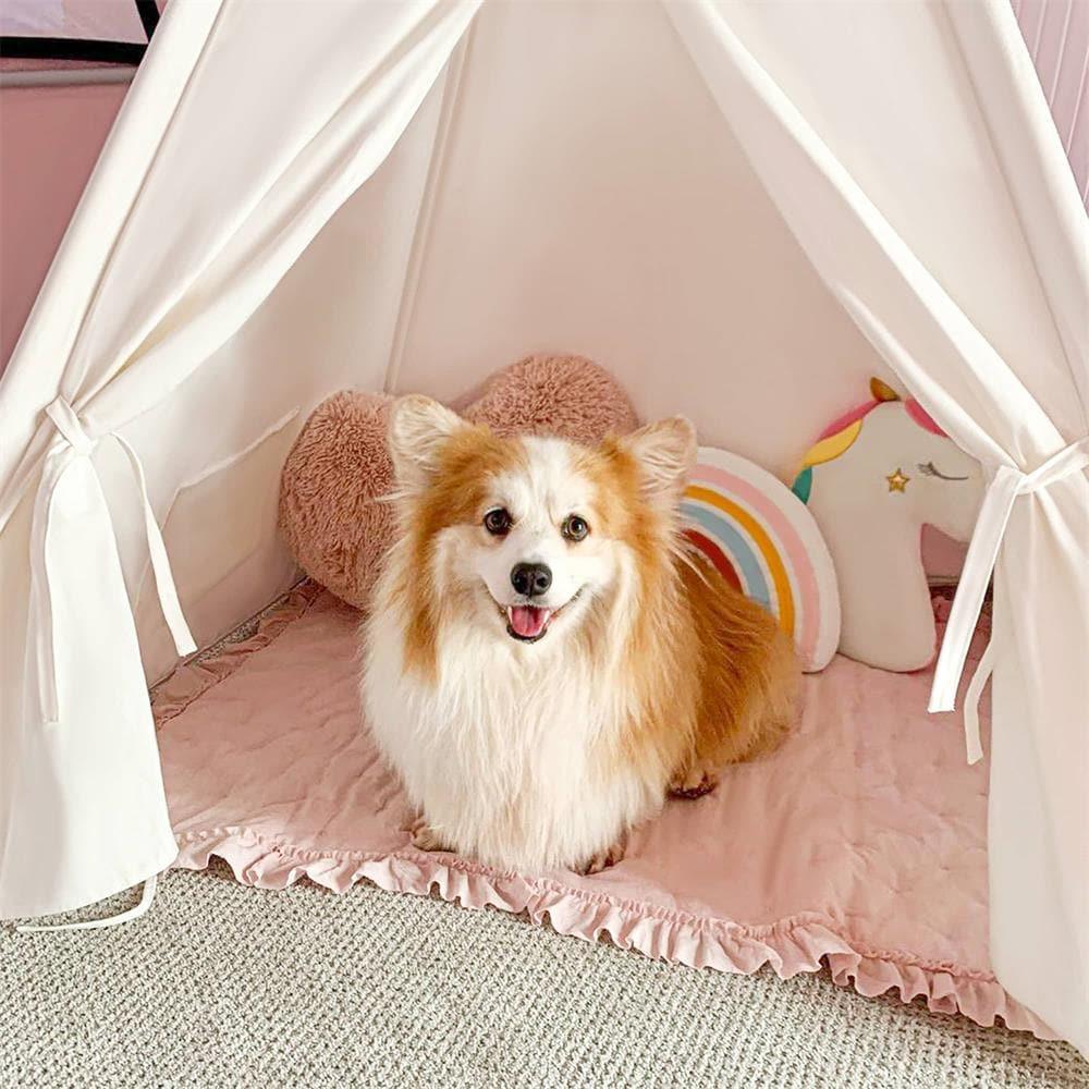A fluffy tan-and-white dog sits happily inside the Tiny Land® Teepee Tent for Kids with Mat by Tiny Land, surrounded by a pink pillow, rainbow cushion, and unicorn pillow on a pink quilted mat.
