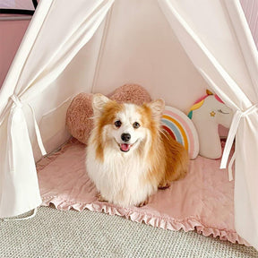 A fluffy tan-and-white dog sits happily inside the Tiny Land® Teepee Tent for Kids with Mat by Tiny Land, surrounded by a pink pillow, rainbow cushion, and unicorn pillow on a pink quilted mat.