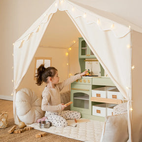A young girl sits inside the Tiny Land® Kids Tent – Large Cozy Tent with Star Lights by Tiny Land, playing with a toy kitchen set and surrounded by soft cushions and wooden toys on a quilted play mat.|fabric
