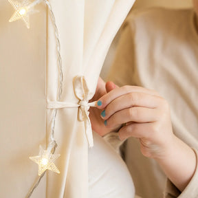 A person with blue nail polish ties a white ribbon around the Tiny Land® Kids Tent – Large Cozy Tent with Star Lights by Tiny Land; star-shaped lights and a quilted play mat nearby create a cozy, softly lit atmosphere.|fabric