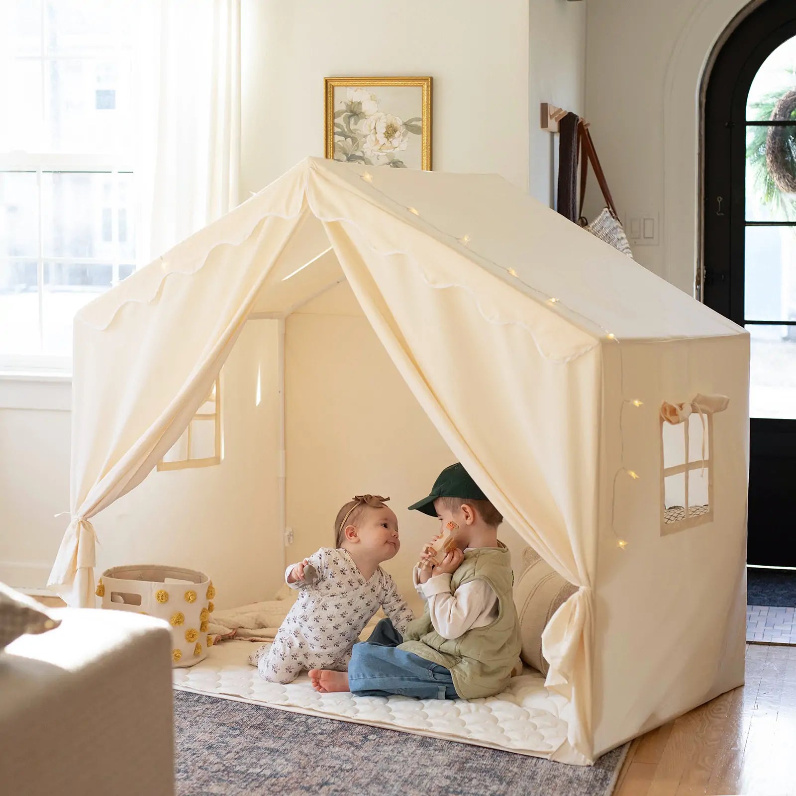 Two children play in a sunlit living room inside the Tiny Land® Kids Tent – Large Cozy Tent with Star Lights. One wears pajamas, the other a cap and vest. A basket and quilted mat add comfort to this inviting scene by Tiny Land.|fabric
