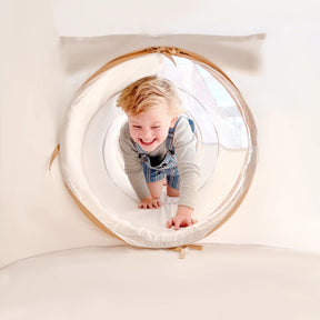 A young child with blonde hair and a grey shirt smiles while crawling in the Tiny Land® Discovery 5-in-1 Play Tunnel (no balls) by Tiny Land, set in a bright, light-colored room.