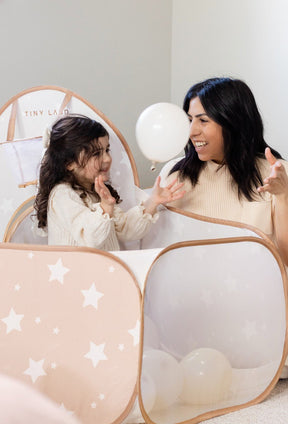 A woman and girl with dark hair smile and play with a white balloon inside the Tiny Land® Compact Adventure 3-in-1 Play Tunnel, surrounded by scattered balloons that turn the tent into their own playful ball pit.|brown