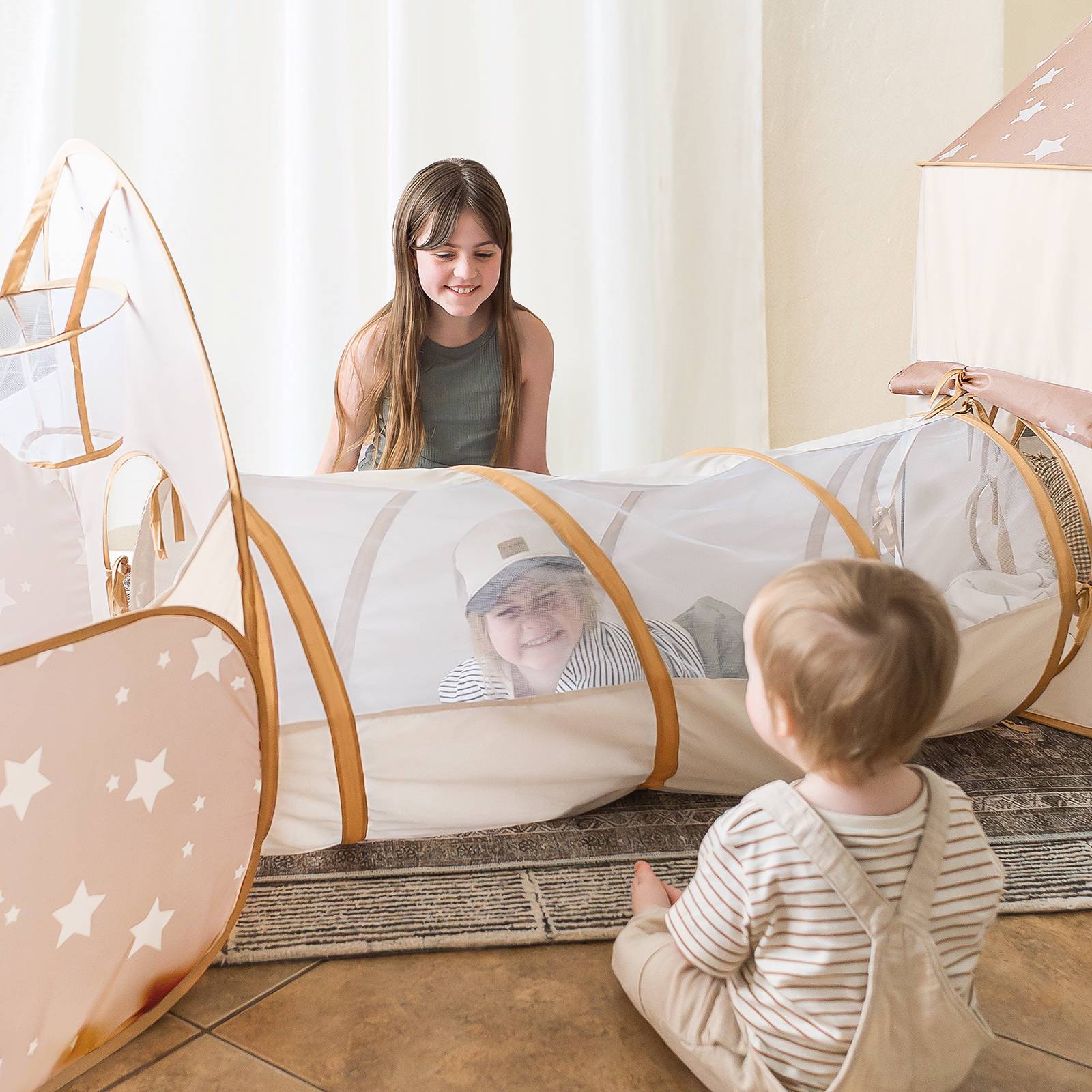 A young girl smiles at two kids playing with the Tiny Land® Compact Adventure 3-in-1 Play Tunnel; one is inside, the other sits at the entrance, transforming their space into an imaginative tent.|brown