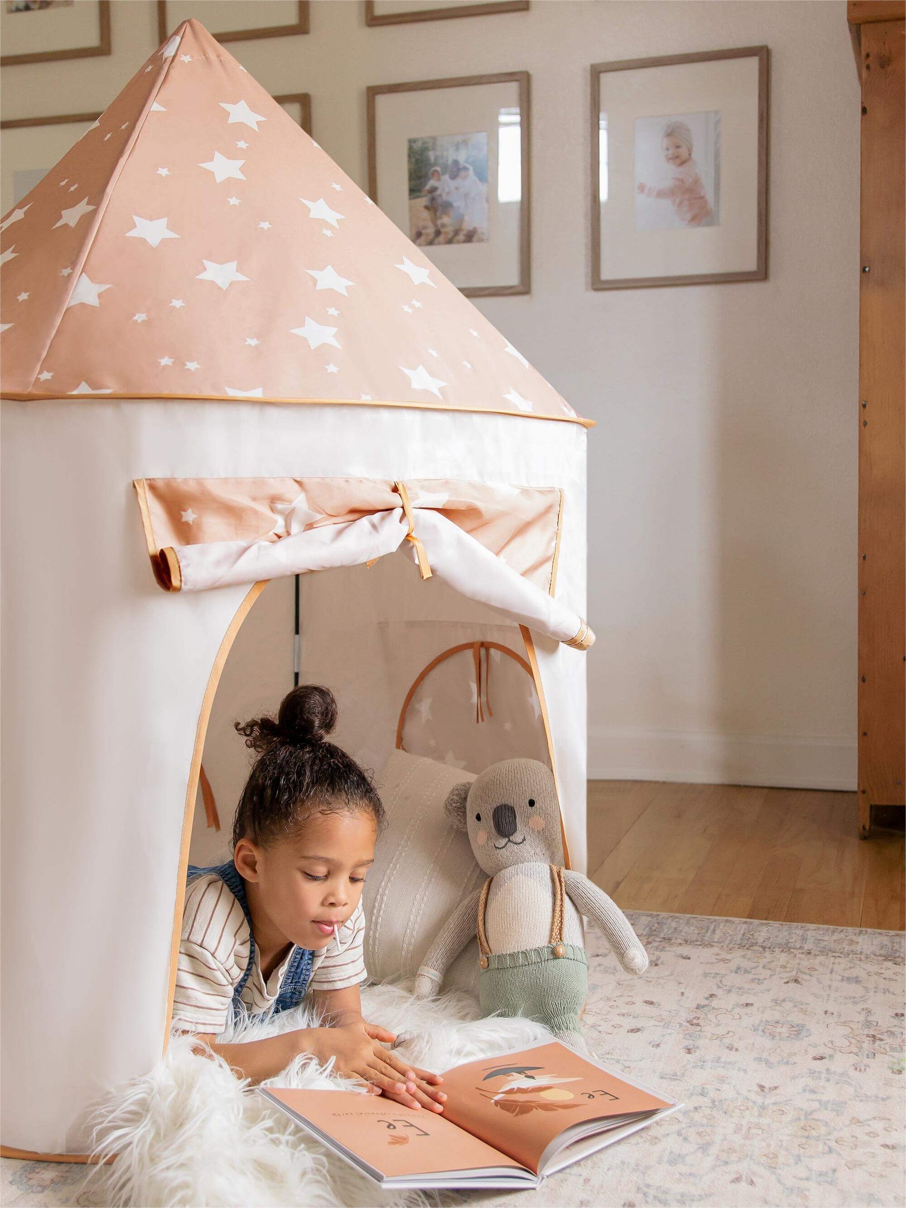 A young girl reads inside the Tiny Land® Adventure 3-in-1 Play Tunnel & Ocean Ball Set by Tiny Land, with a starry play tent, a stuffed animal on a cozy rug, and framed photos warming the room.|brown