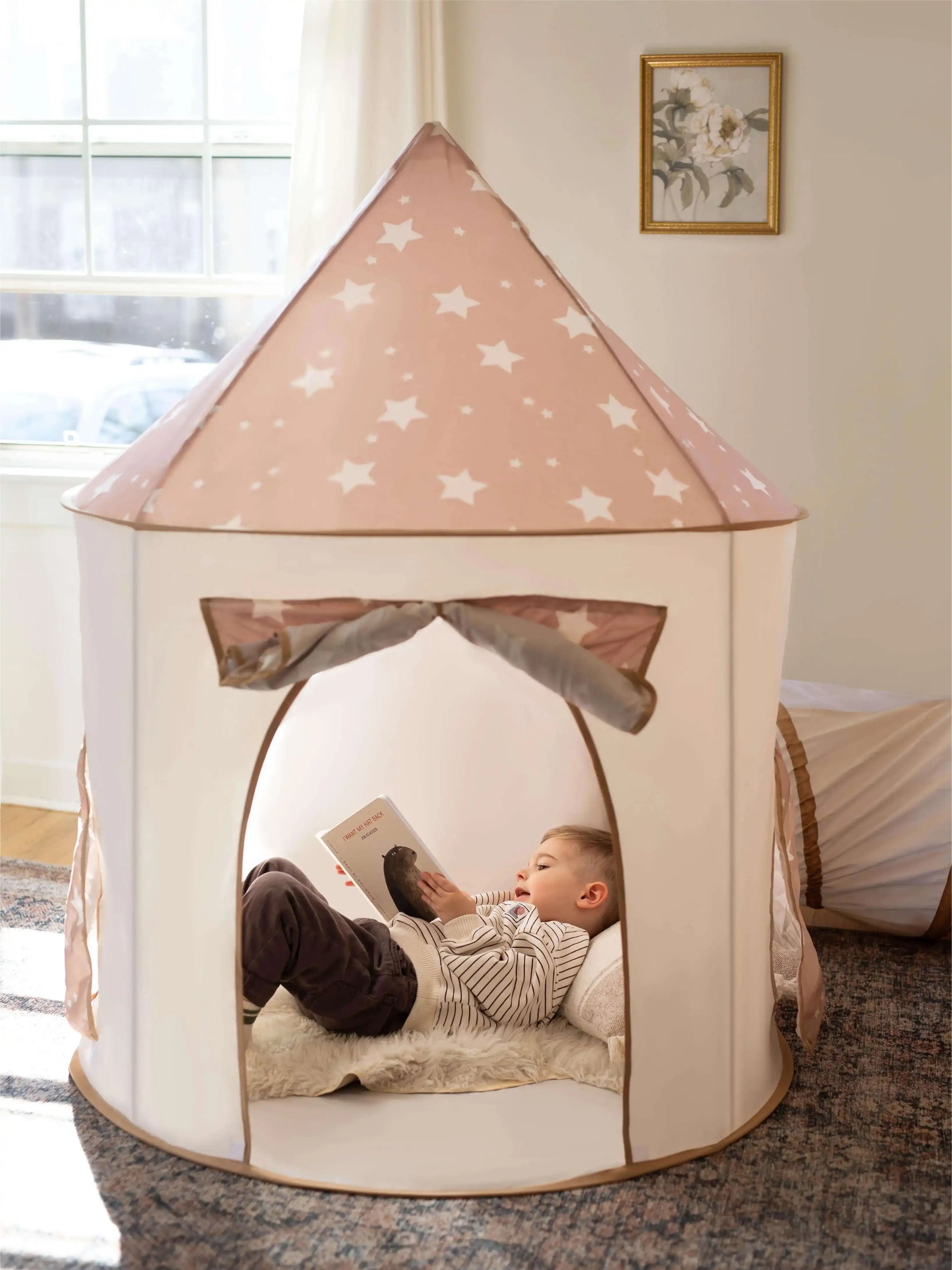 A young child relaxes inside the Tiny Land® Compact Adventure 3-in-1 Play Tunnel, reading a book. Soft light filters in, highlighting the playful space by Tiny Land, with a cozy atmosphere and a framed picture on the wall.|brown