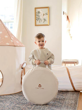 A young child smiles indoors, holding a Tiny Land storage bag. Behind them are the Tiny Land® Adventure 3-in-1 Play Tunnel & Ocean Ball Set, featuring a star-patterned tent, cozy ball pit, and soft play area in a bright room.|brown