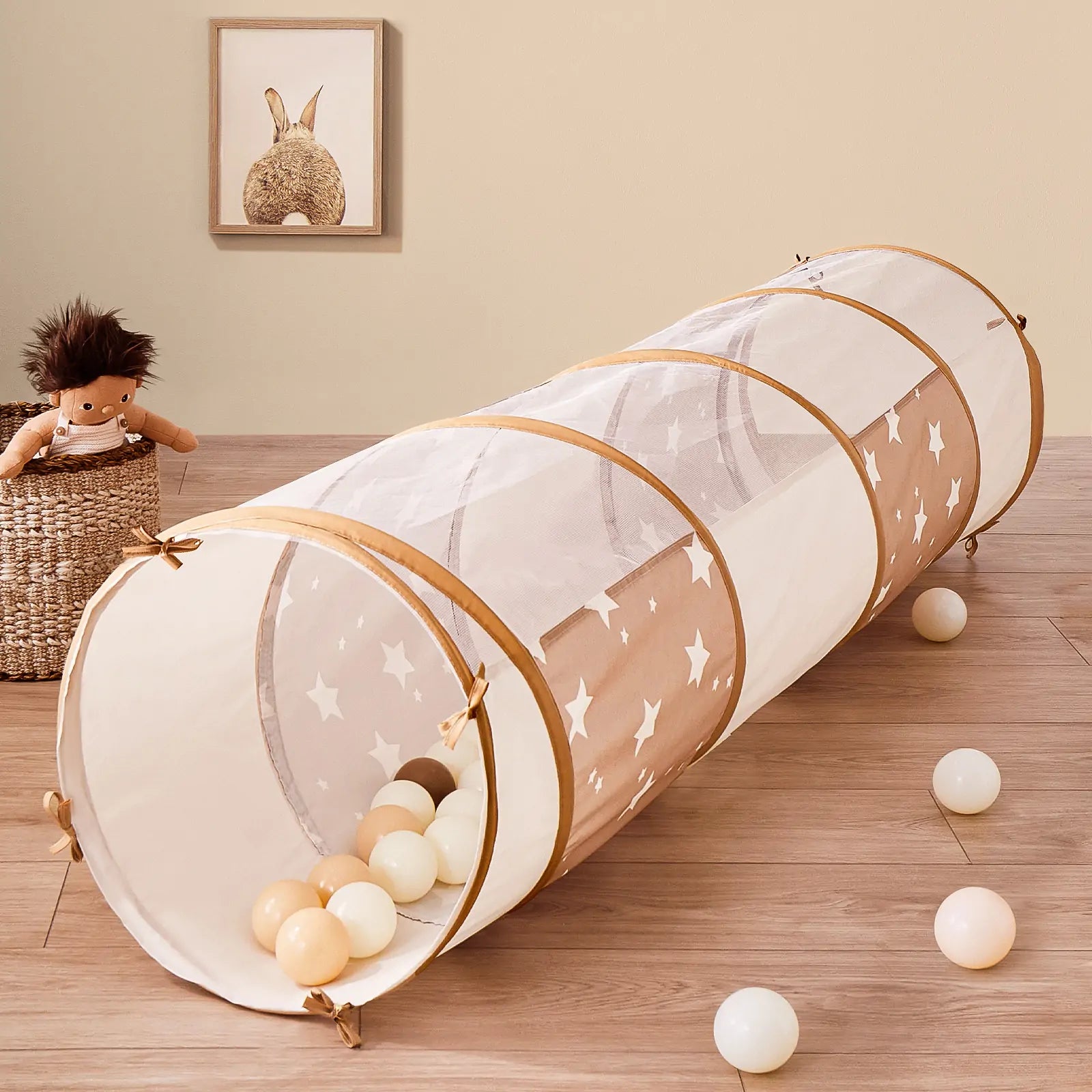 The Tiny Land® Violet PlayNest Climbing Ball Pit Set with Tunnel sits on a wooden floor, filled with colorful foam ball pit balls. Nearby, a doll rests in a woven basket while framed animal artwork hangs on the beige wall in the background, creating a cozy indoor playground atmosphere.|Pink