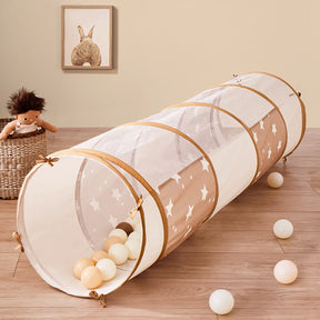 The Tiny Land® Violet PlayNest Climbing Ball Pit Set with Tunnel sits on a wooden floor, filled with colorful foam ball pit balls. Nearby, a doll rests in a woven basket while framed animal artwork hangs on the beige wall in the background, creating a cozy indoor playground atmosphere.|Pink