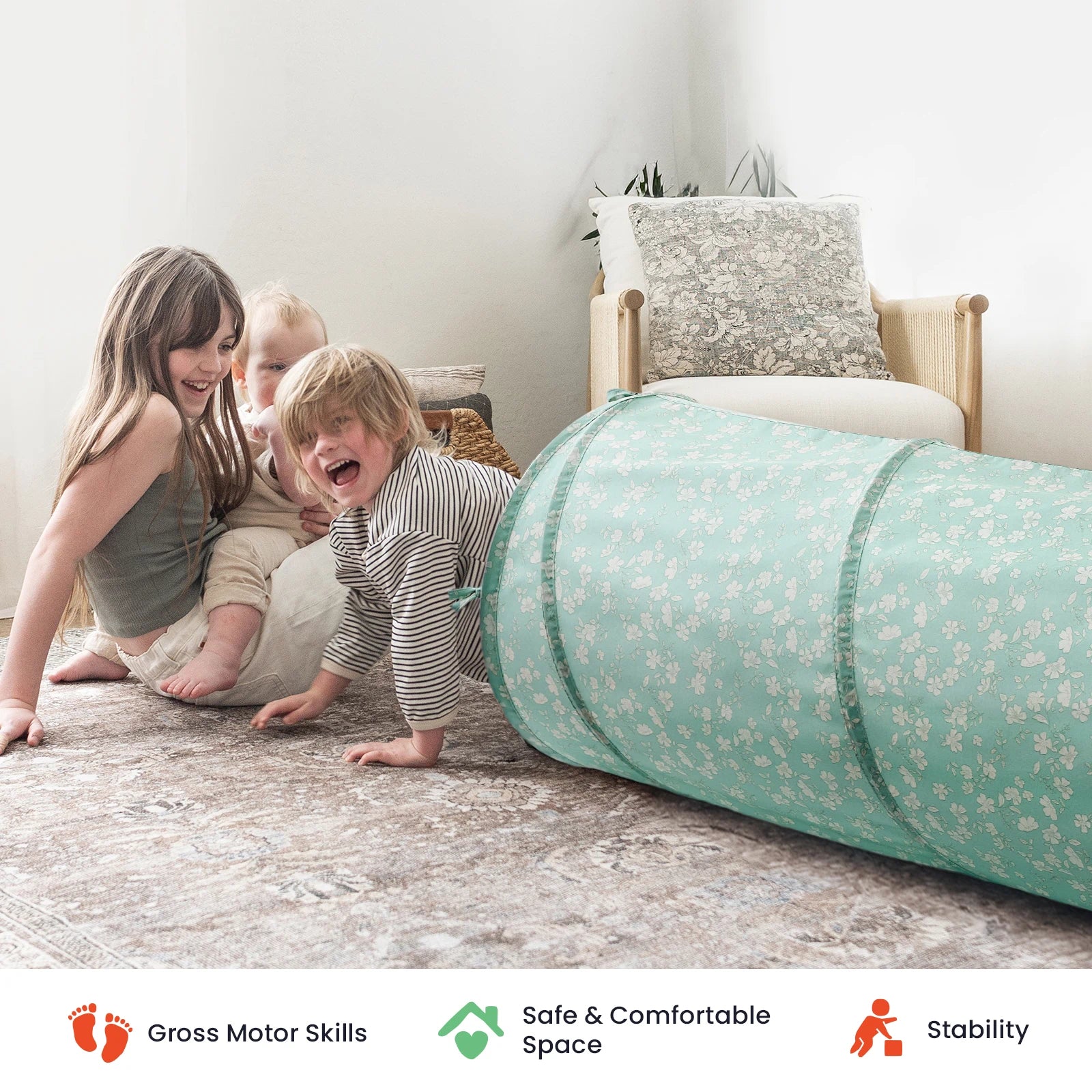 Three young children play and crawl through the Tiny Land® Adventure Play Tunnel, a light blue floral tunnel designed by Tiny Land, in a cozy, softly lit living room featuring a beige armchair and cushions in the background. Icons below highlight motor skills development, safety, and stability.|Green