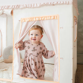 A young child with light hair and a floral dress smiles while holding open the pink curtains of the Tiny Land® Sweet Citrus Kids Play Tent – a white and pink indoor playhouse by Tiny Land, perfect for creative play.|pink