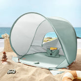 The Tiny Land® Baby Beach Tent—a compact, sea blue play tent by Tiny Land—sits on sandy shore near the sea with a yellow book and toys inside. Nearby are a woven basket, stacked stones, swim ring, and the ocean in the background.|Sea Blue