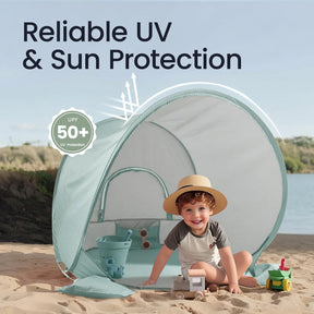 A young child smiles inside the Tiny Land® Baby Beach Tent – Sea Blue by Tiny Land on a sandy beach, surrounded by toys. The open pop-up tent features illustrated sun rays and “Reliable UV & Sun Protection UPF 50+” text above.|Sea Blue