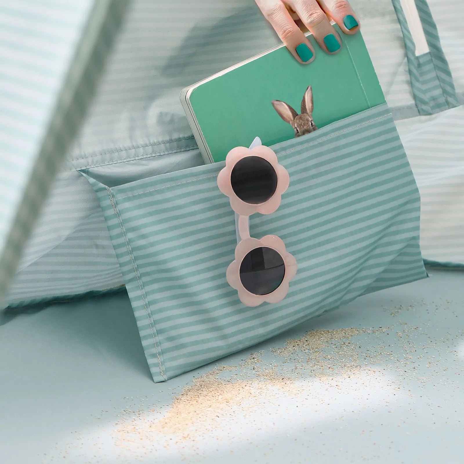 A hand with teal nails holds a green notebook featuring a rabbit, partially tucked into the striped pocket of the Tiny Land® Baby Beach Tent – Sea Blue by Tiny Land, with sand nearby and flower-shaped sunglasses hanging from the pocket.|Sea Blue