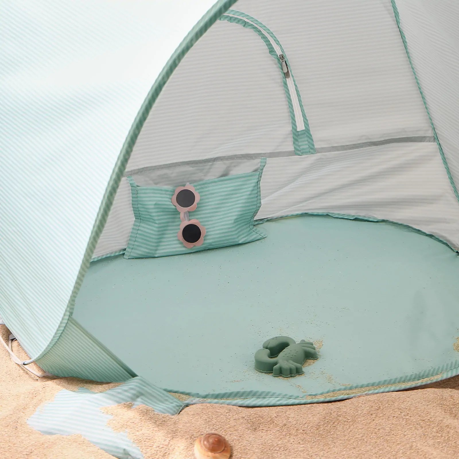 The Tiny Land® Baby Beach Tent – Sea Blue Play Tent by Tiny Land sits on the sand. Inside are a matching striped pillow with pink sunglasses on top and a green turtle-shaped toy, with some sand scattered on the tent floor.|Sea Blue