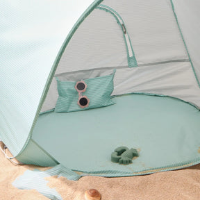 The Tiny Land® Baby Beach Tent – Sea Blue Play Tent by Tiny Land sits on the sand. Inside are a matching striped pillow with pink sunglasses on top and a green turtle-shaped toy, with some sand scattered on the tent floor.|Sea Blue