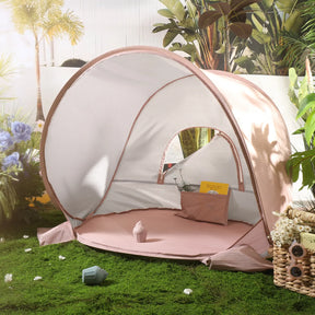 A Tiny Land® Baby Beach Tent in blush pink is set up on grass amid flowers. Inside the tent is a small pillow and yellow envelope, with a toy and flower basket nearby. Sunlight softly brightens this cozy outdoor scene.|Blush Pink