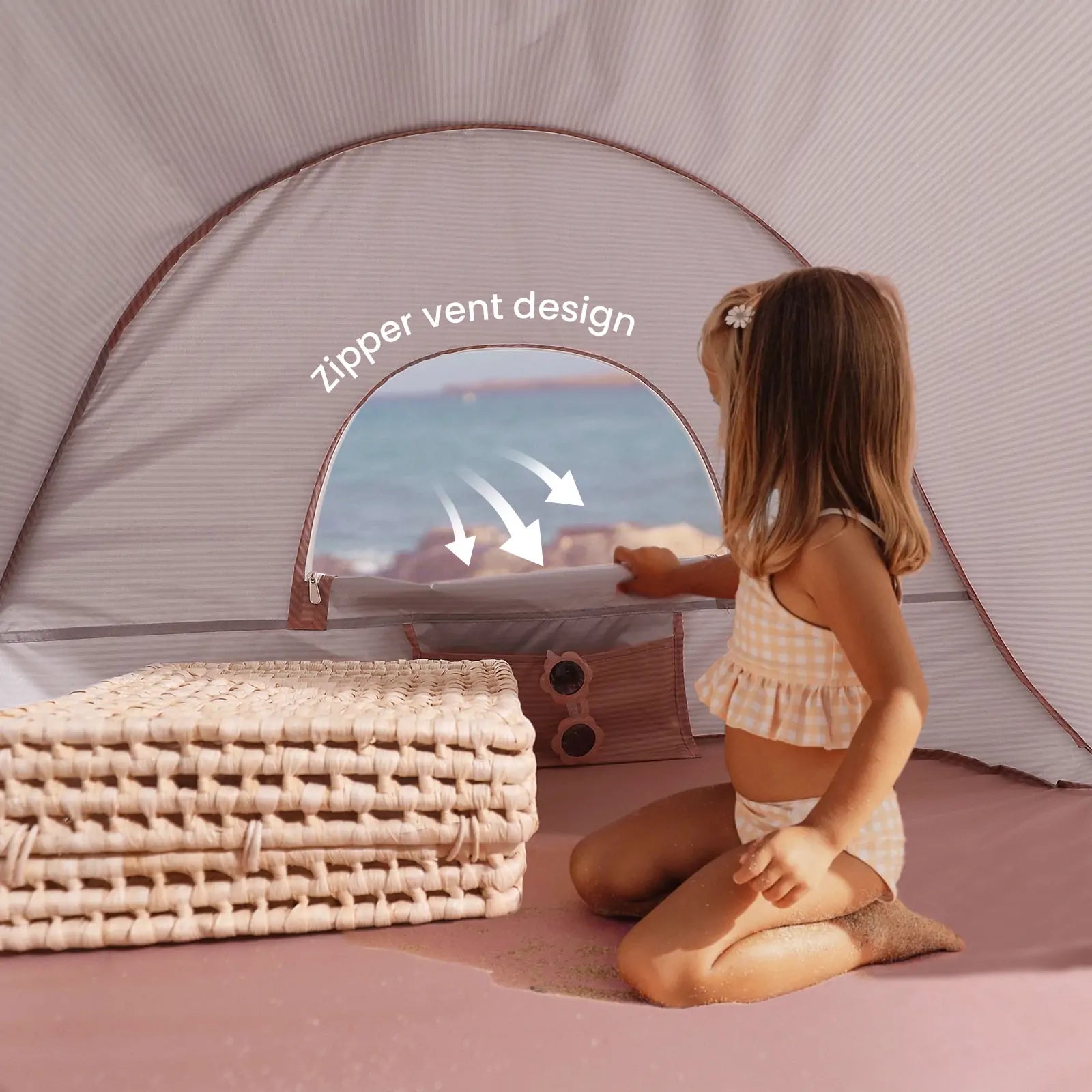 A young girl in a checked swimsuit enjoys the Tiny Land® Baby Beach Tent in blush pink on the sand, opening its zippered vent. Folded mats sit nearby, with the sea visible through the window. “Zipper vent design” is displayed in white text.|Blush Pink