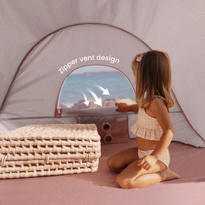A young girl in a checked swimsuit enjoys the Tiny Land® Baby Beach Tent in blush pink on the sand, opening its zippered vent. Folded mats sit nearby, with the sea visible through the window. “Zipper vent design” is displayed in white text.|Blush Pink