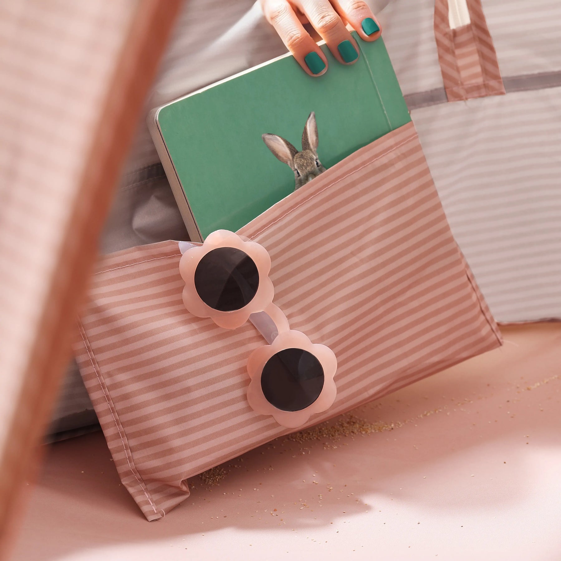 A hand packs a green notebook with a rabbit cover into a pink-and-white pouch alongside flower-shaped sunglasses—ideal for unwinding by the Tiny Land® Baby Beach Tent – Blush Pink Play Tent. Sand is sprinkled across the light pink surface.|Blush Pink