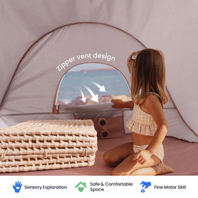 A young girl in a checkered swimsuit sits inside the Tiny Land® Baby Beach Tent – Play Tent for Outdoor Fun by Tiny Land, which features a convenient zipper vent and is set up near woven mats. This beach tent provides sensory exploration, a safe space, UV protection, and supports fine motor skill development.|Blush Pink