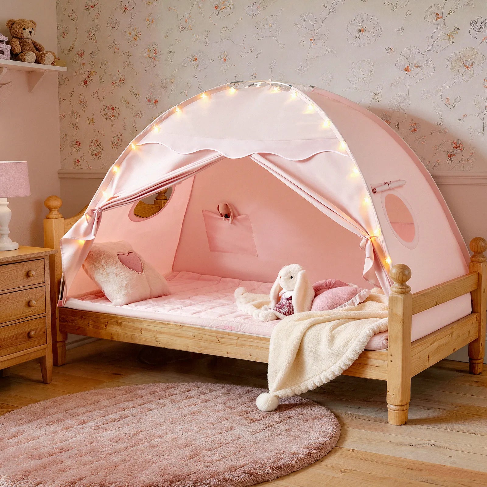 The Tiny Land® Versatile Kids Bed Tent & Play Tent by Tiny Land features a cozy wooden-framed children’s bed with a pink canopy, creating the perfect indoor tent for kids. It’s beautifully accented with fairy lights, a plush bunny, heart-shaped pillow, soft blanket, round rug, and floral wallpaper in a softly lit room.