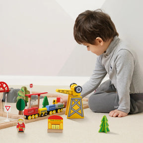 A young boy in grey pajamas plays on the floor with the Tiny Land® Wooden Track Trains 55 Pcs set by Tiny Land, featuring an interactive crane, tracks, trees, a train station, and small figurines.|55 Pcs