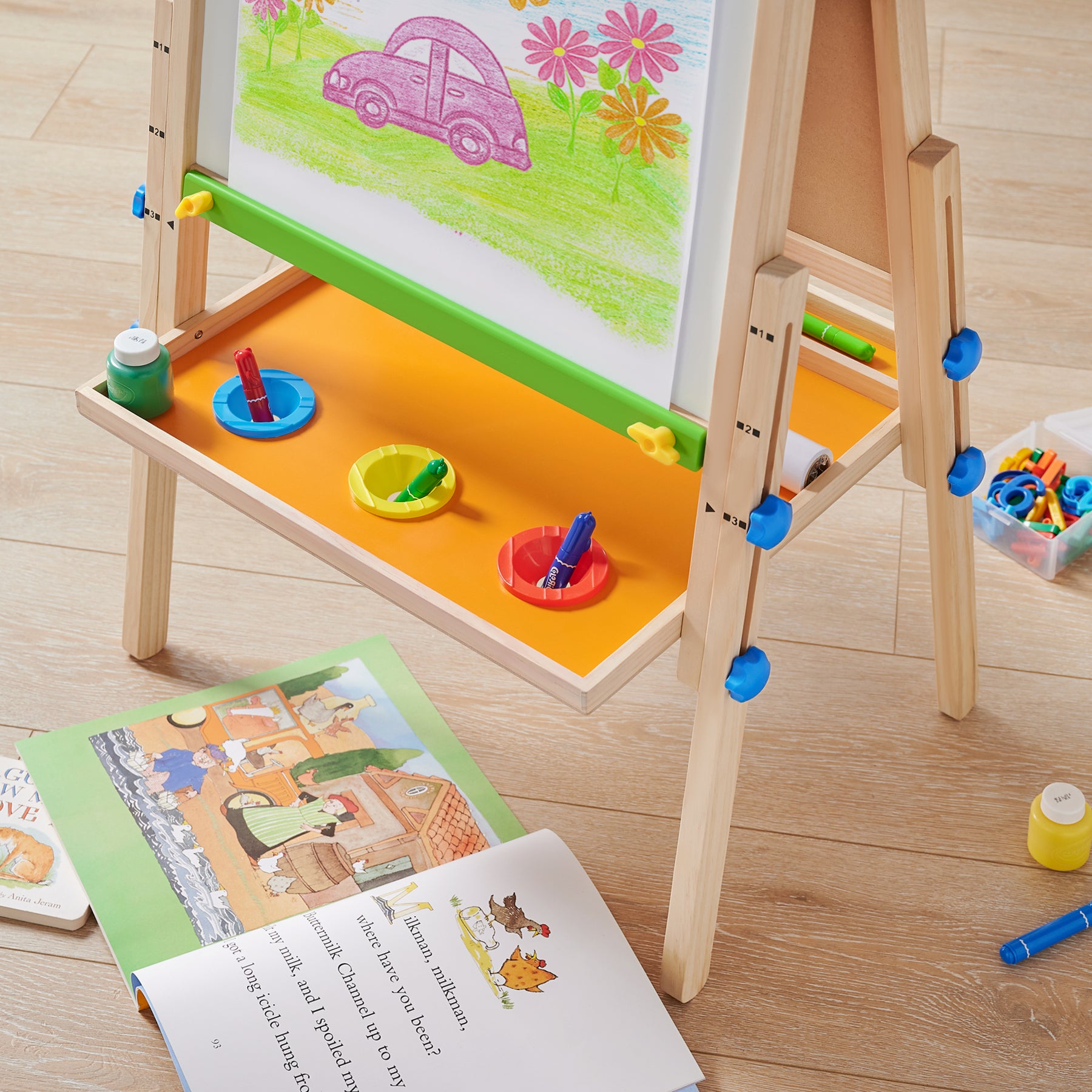 The Tiny Land® 3-in-1 Art Easel & Paper Roll Set displays a drawing of a purple car and flowers. Paint pots, markers, and brushes from the set are on the shelf, while children’s books lay scattered on the wooden floor nearby.|red