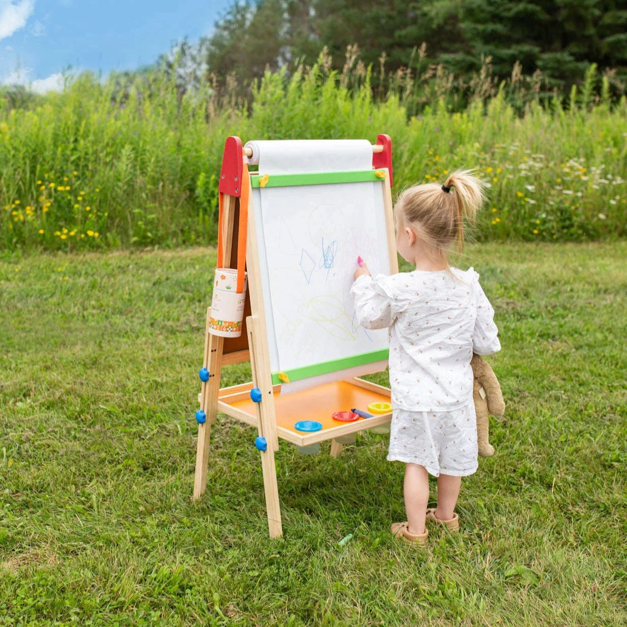 A young child in light pajamas holds a stuffed animal and draws on the Tiny Land® 3-in-1 Art Easel & Paper Roll Set in a grassy field, surrounded by wildflowers and trees, enjoying a peaceful moment of creativity.|red