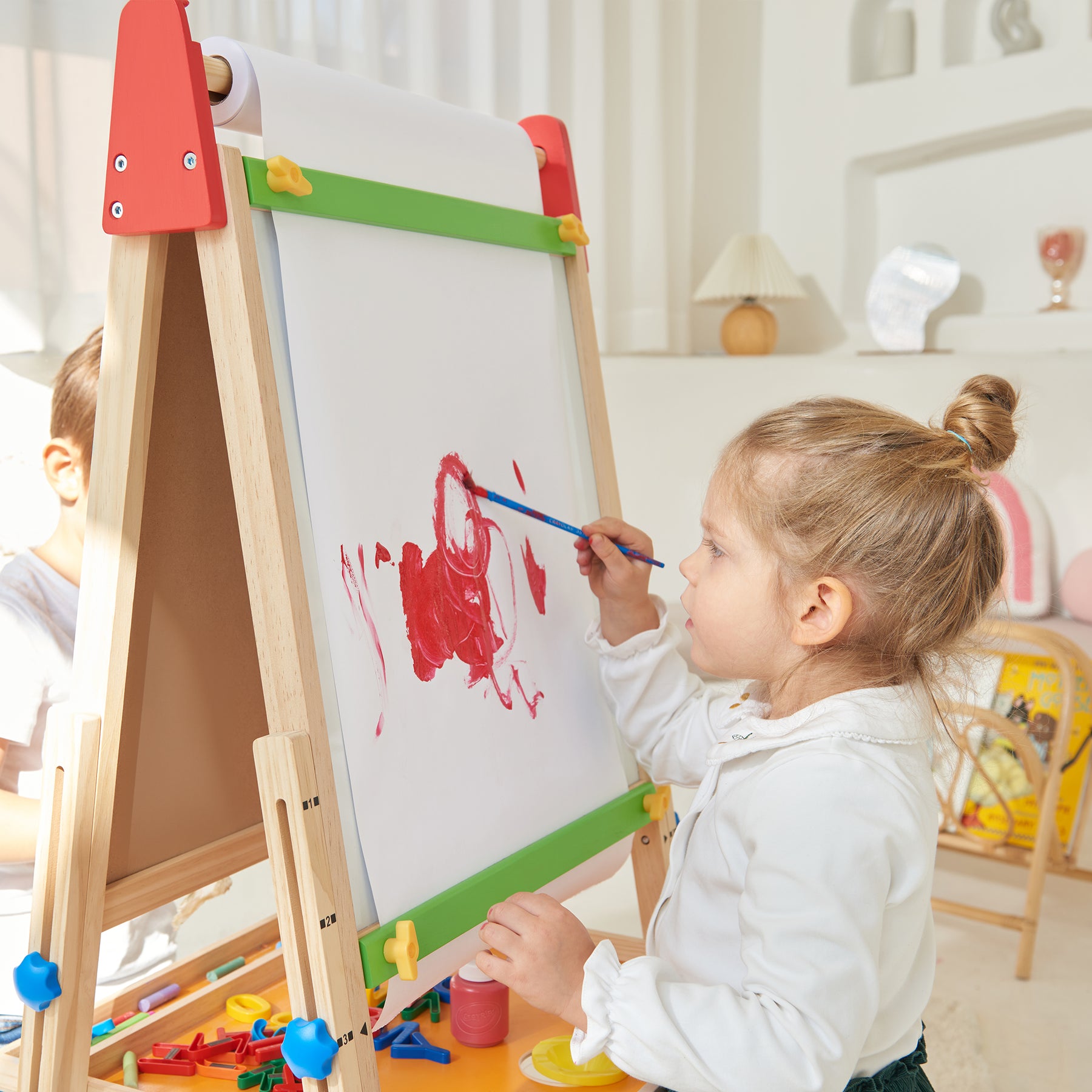A young girl with light brown hair in a ponytail paints with red on the Tiny Land® 3-in-1 Art Easel & Paper Roll Set by Tiny Land in a bright, cozy room, with art supplies scattered nearby.|red