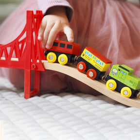 A child in a pink top and skirt pushes colorful trains over a red bridge from the Tiny Land® Wooden Train Set 110 Pcs by Tiny Land, compatible with all major brands, on a quilted surface.|110 Pcs