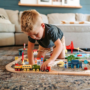 A young boy plays on the floor with the Tiny Land® Wooden Train Set 110 Pcs, compatible with all brands, surrounded by miniature buildings, signs, and tracks in a cozy living room with a sofa in the background.|110 Pcs