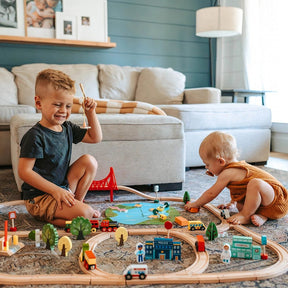 Two young children play on a light-colored rug with the Tiny Land® Wooden Train Set 110 Pcs. The older child smiles while holding a train, as the younger explores tracks and toys compatible with all brands from Tiny Land.|110 Pcs