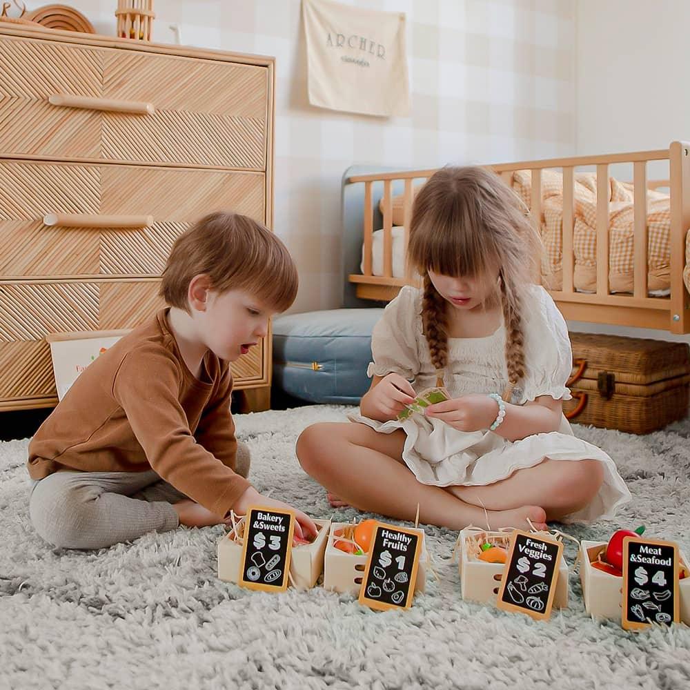 Two young children sit on a soft carpet, playing with the Tiny Land Play Food Wooden Food for Play Kitchen & Wooden Rack set, with baskets labeled “Bakery Sweets,” “Healthy Fruit,” “Fruit & Veggies,” and “Meat & Seafood” in a cozy bedroom.
