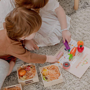 Two kids sit on a carpet, playing with the Tiny Land® Wooden Food for Play Kitchen & Wooden Rack by Tiny Land Play Food. They arrange realistic wooden play food in crates and point to a Farmers Market menu on the floor.